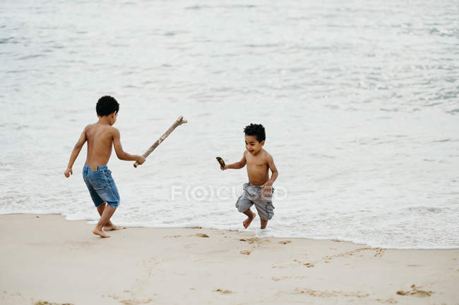 Zwei lustige afrikanisch-amerikanische Brüder mit Stöcken spielen zusammen am Sandstrand in der Nähe des Meeres — Stockfoto