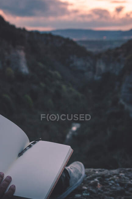 View to small river in canyon and legs of person with book sitting on the edge — Stock Photo