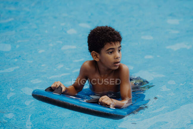 Adorable chico afroamericano con flotador mirando hacia otro lado mientras nada en agua limpia de la piscina - foto de stock