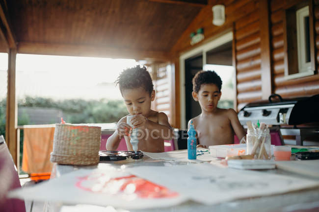 Deux garçons afro-américains torse nu utilisant une peinture brillante pour faire des images abstraites sur la table à la maison — Photo de stock