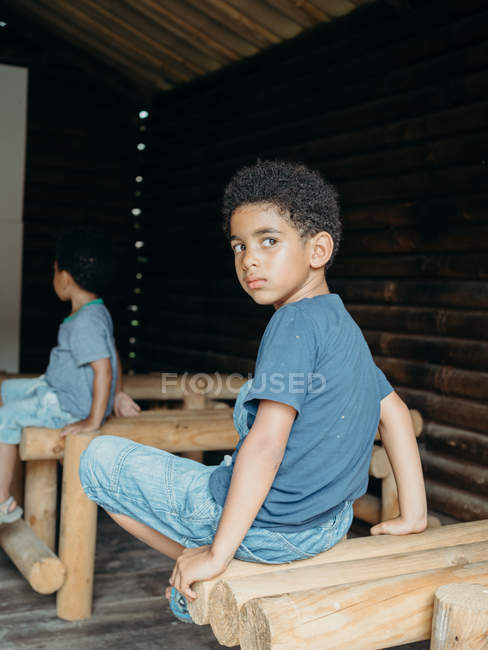 Cute African American boy in casual outfit looking over shoulder while sitting on wooden structure inside lumber building — Stock Photo