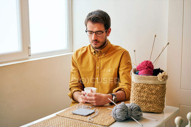 Дорослий чоловік насолоджується свіжим гарячим чаєм і дивиться вбік, сидячи за столом біля в'язаних голок і пряжі — стокове фото
