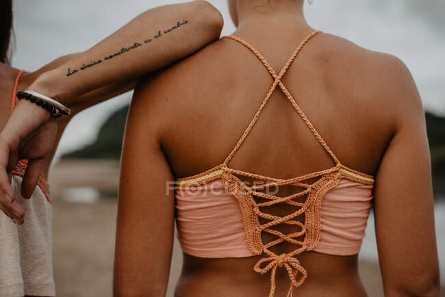 Cortar hembra con el brazo tatuado apoyado en el hombro de un amigo irreconocible mientras pasan tiempo en la playa juntos - foto de stock