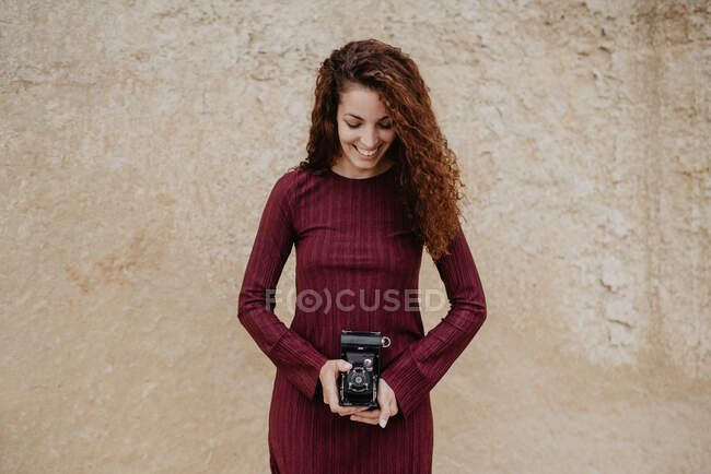 Beautiful female in stylish dress smiling and holding retro camera while standing near shabby wall — Stock Photo