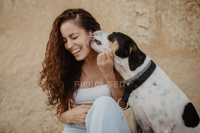 Funny dog licking cheek of excited young lady against weathered building wall on street — Stock Photo
