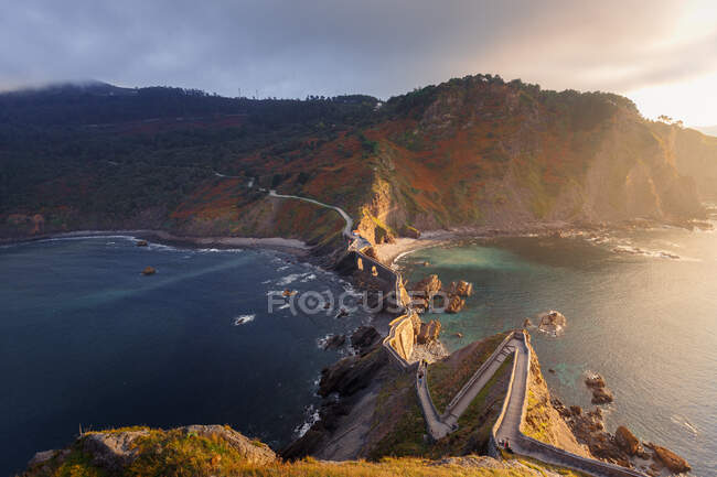 Desde arriba pintoresco paisaje de la isla Gaztelugatxe con puente de piedra largo que pasa a través de la orilla del mar en el día ventoso - foto de stock