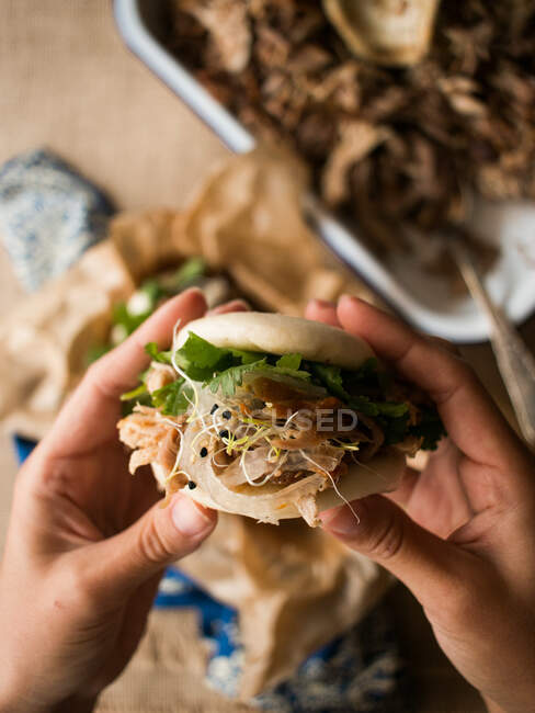 Mãos de pessoa anônima segurando pão de bao fresco com saboroso porco puxado e ervas sobre a mesa na cozinha — Fotografia de Stock