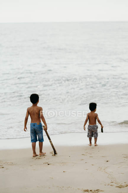 Два смешных афроамериканских брата с палками играют вместе на песчаном берегу у моря — стоковое фото