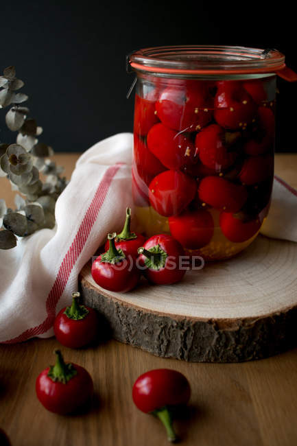 Still life with jar of tasty pickled pimientos peppers placed on piece of wood near fabric napkin and plant sprig. — Stock Photo