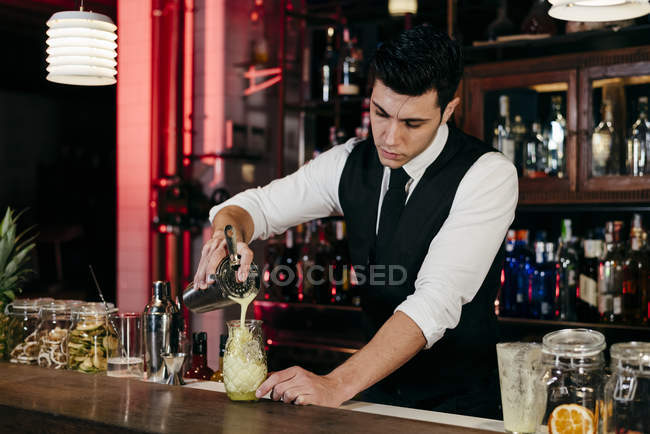 Young elegant barman working behind a bar counter pouring drink from shaker to a glass — Stock Photo