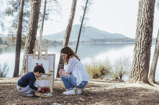 Vista lateral da mãe olhando para a pintura do menino bonito no cavalete enquanto passar o tempo perto do lago no campo juntos — Fotografia de Stock