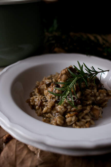 Тарелка вкусного рисового ризотто с мясом кролика и грибами, украшенная свежей веточкой розмарина на кухне — стоковое фото