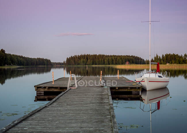 Pequeno iate branco com velas para baixo no cais de madeira à beira do lago perto de uma pequena aldeia na Finlândia — Fotografia de Stock