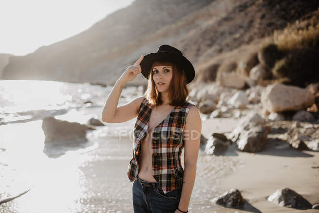 Attractive woman with unbuttoned checkered shirt posing near sea water on rocky coast against mountains on sunny day in countryside — Stock Photo