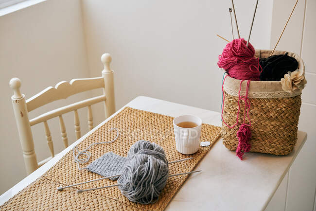 Cup of warm tea placed on table near basket with knitting yarn and needles in cozy room — Stock Photo
