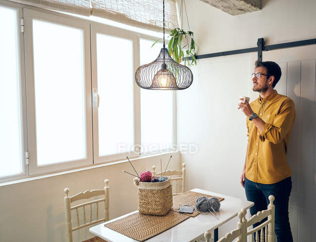 Взрослый мужчина наслаждается горячим чаем и смотрит в сторону, сидя за столом возле вязальных спиц и пряжи — стоковое фото