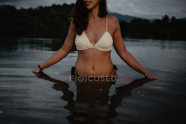 Неузнаваемая молодая женщина в купальниках, стоящая в спокойной воде пруда вечером на природе — стоковое фото