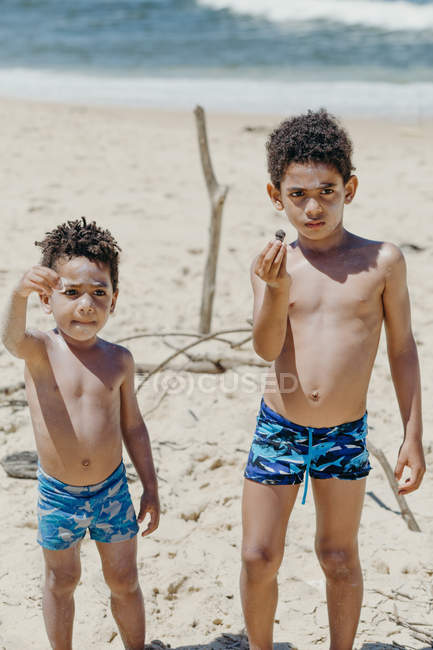 Сладкие афроамериканские дети смотрят в камеру и демонстрируют крошечную раковину, стоя у синего моря. — стоковое фото