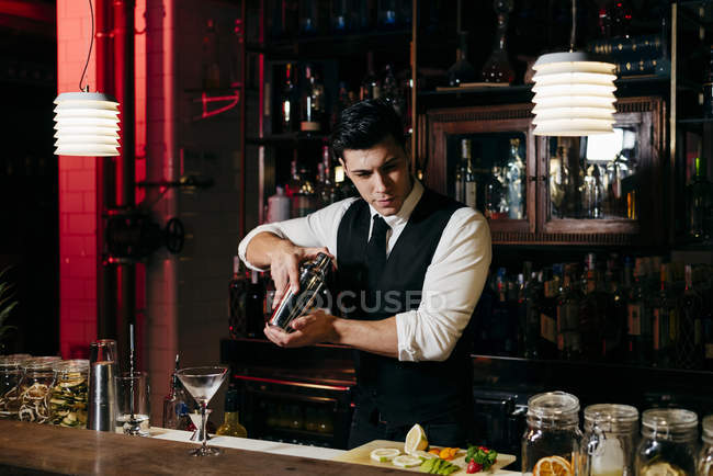 Jovem barman elegante trabalhando atrás de um balcão de bar misturando bebidas em uma coqueteleira — Fotografia de Stock