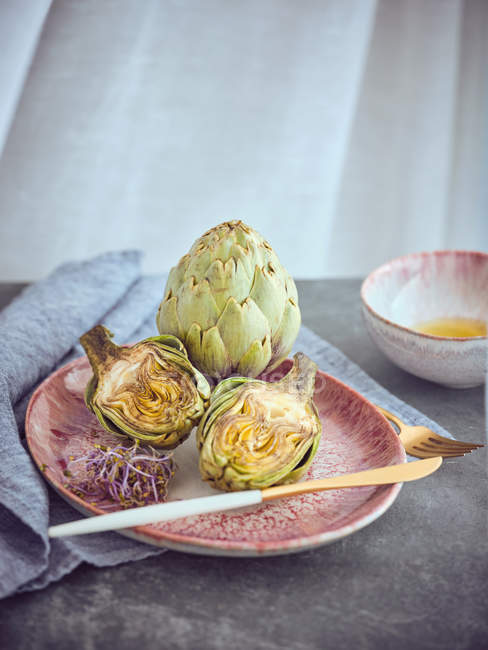 Whole and halved fresh artichokes and sprouts on pink ceramic plate on table — Stock Photo