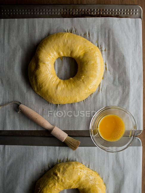 Bowl with fresh egg yolk placed on lumber table near delicious uncooked Rosca de Reyes pastry. — Stock Photo