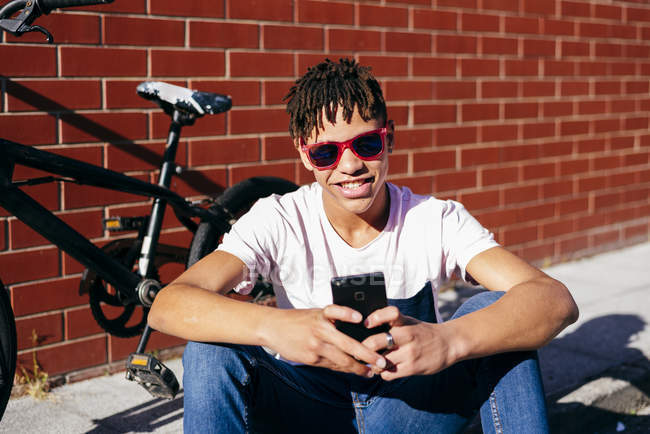 Joven hombre negro feliz navegando teléfono inteligente cerca de la bicicleta - foto de stock