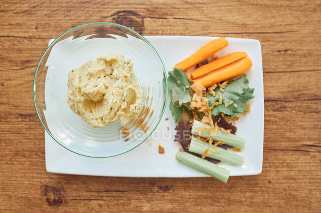 De dessus disposition des légumes naturels sains comme la carotte, concombre, salade et bol de houmous savoureux sur assiette blanche — Photo de stock