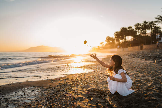 Niña en vestido blanco sentado en la playa y jugando con piedras en el fondo de la puesta del sol - foto de stock