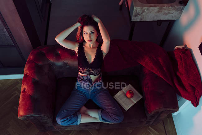 Joven mujer sexy sentada en el sofá con manzana y libro - foto de stock