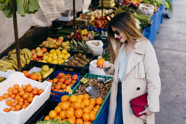 Joven feliz bonita mujer en ropa elegante y gafas de sol elegir frutas en el mercado en la calle - foto de stock