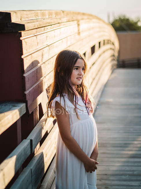 Portrait of cute little girl in white dress standing near wooden wall in sunlight — Stock Photo