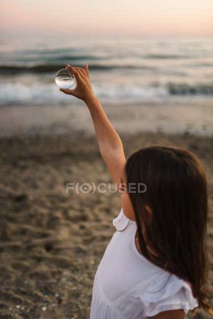 Little female child with brown hair holding glass ball on background of waves — Stock Photo