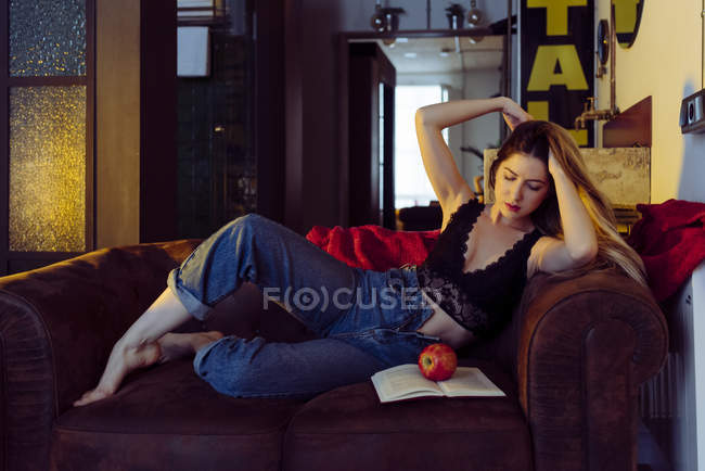 Giovane donna sexy seduta sul divano con mela e libro — Foto stock
