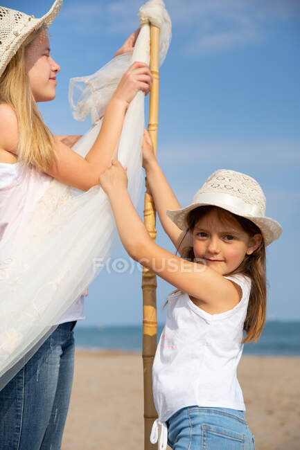 Девушки в шляпах прикрепляют навес на шест на песке на пляже — стоковое фото
