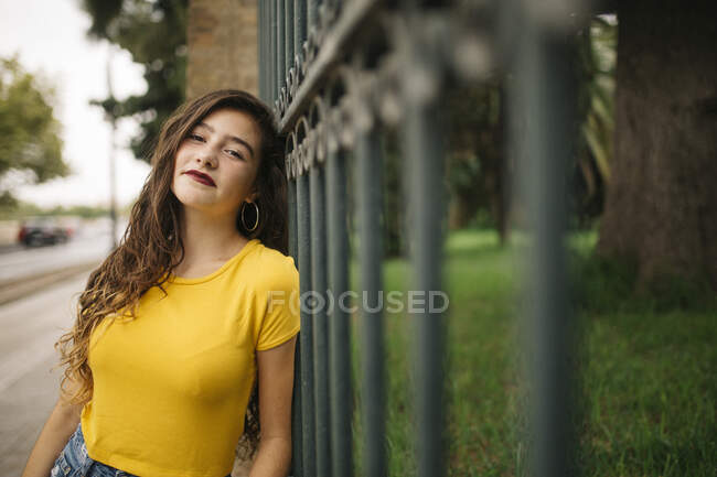 Satisfeito jovem fêmea em roupa casual sorrindo e olhando para a câmera enquanto se inclina na cerca do parque da cidade — Fotografia de Stock