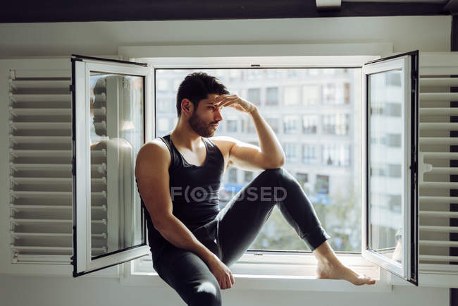 Jovem macho bem sucedido em singlet casual sentado na janela e olhando para longe — Fotografia de Stock