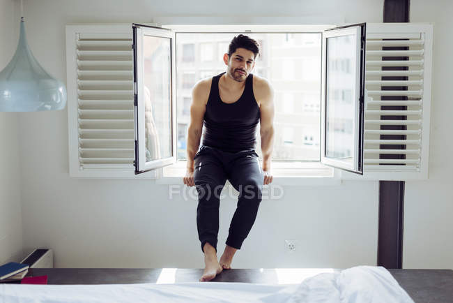 Young cheerful successful male in casual singlet sitting on window near bed with books and looking at camera — Stock Photo
