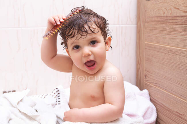 Adorable bebé mirando a la cámara y peinando el cabello mojado mientras está sentado en la toalla en el baño después de la ducha - foto de stock