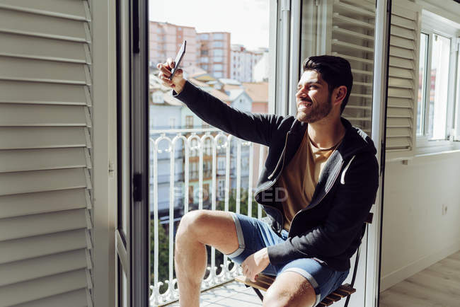 Side view of young joyful successful man in casual apparel sitting on chair near balcony and taking selfie with smartphone — Stock Photo