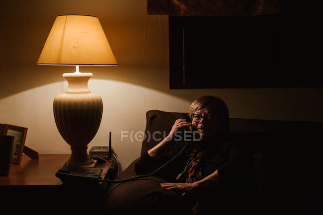 Радостная пожилая женщина улыбается и отвечает на телефонный звонок, сидя в темной комнате вечером дома — стоковое фото