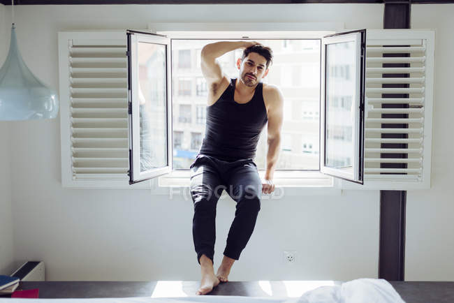 Молодой веселый успешный мужчина в случайном сингле сидит на окне рядом с кроватью с книгами и смотрит в камеру — стоковое фото