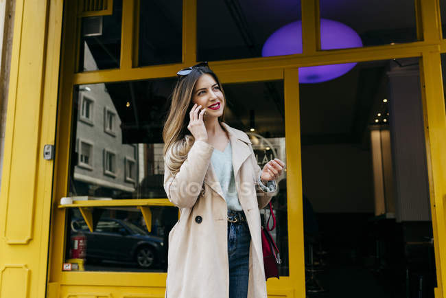 Молода весела жінка стоїть біля кафе і розмовляє по смартфонах — стокове фото