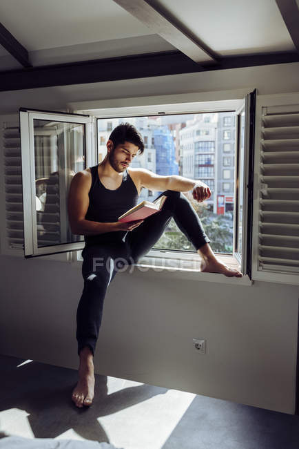 Вид збоку молодого серйозного красивого чоловіка в повсякденній без рукавів сорочці, що сидить на вікні і читає цікаву книгу — стокове фото