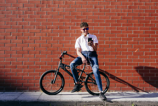 Молодой улыбающийся афроамериканец с афроволосами в повседневной одежде сидит на велосипеде и пользуется смартфоном — стоковое фото