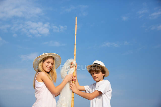 Kinder mit Hüten befestigen Sonnensegel an Stange auf Sand am Strand — Stockfoto