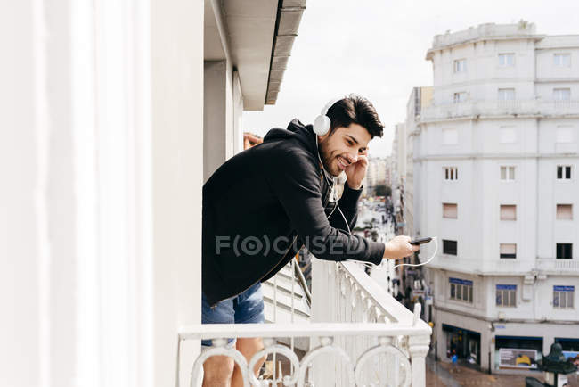 Joven sonriente guapo hombre casual en auriculares apoyados en la barandilla de la terraza con teléfono inteligente y mirando hacia otro lado - foto de stock