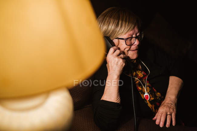 Mulher idosa feliz sorrindo e respondendo telefonema enquanto sentado no quarto escuro à noite em casa — Fotografia de Stock