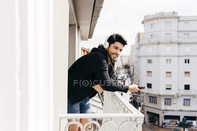 Молодий усміхнений красивий випадковий чоловік у навушниках спирається на тераси поручні з смартфоном і дивитися геть — стокове фото