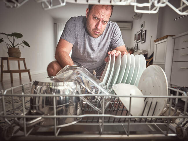 Недовольный взрослый мужчина кладет тарелку в посудомоечную машину на стильной кухне дома — стоковое фото