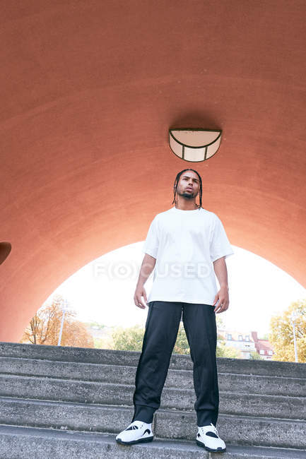 Confident African American male in casual outfit standing on steps near modern arch on city street — Stock Photo
